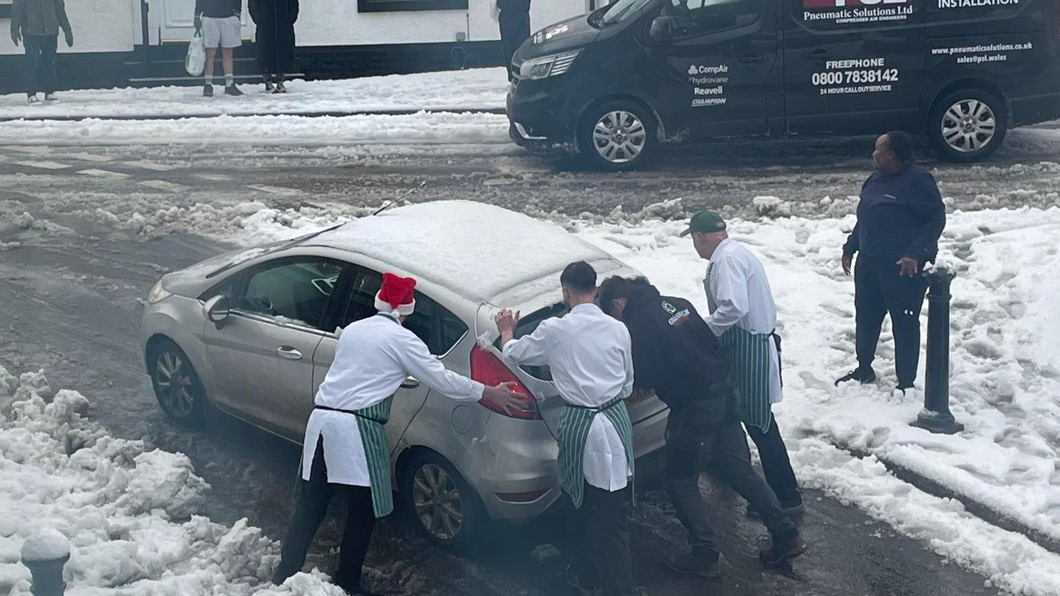 Men in white shirts and green aprons and santa hats  pushing car in snow 