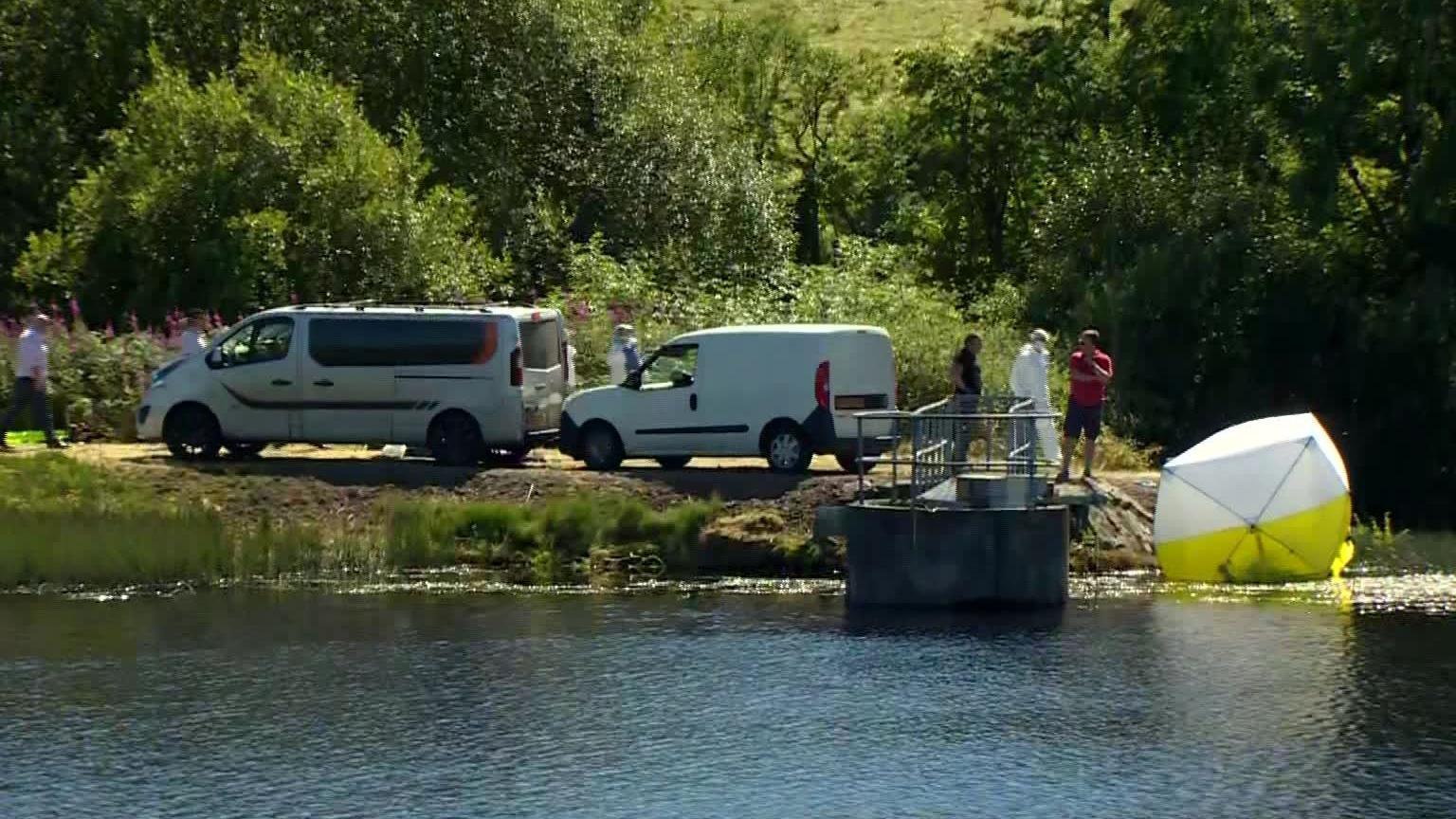 An image of the banks of a reservoir. There are two white vans, and severel people standing around. One of them is in white forensic overalls and there is a forensics white and yellow tent at the water's edge. 