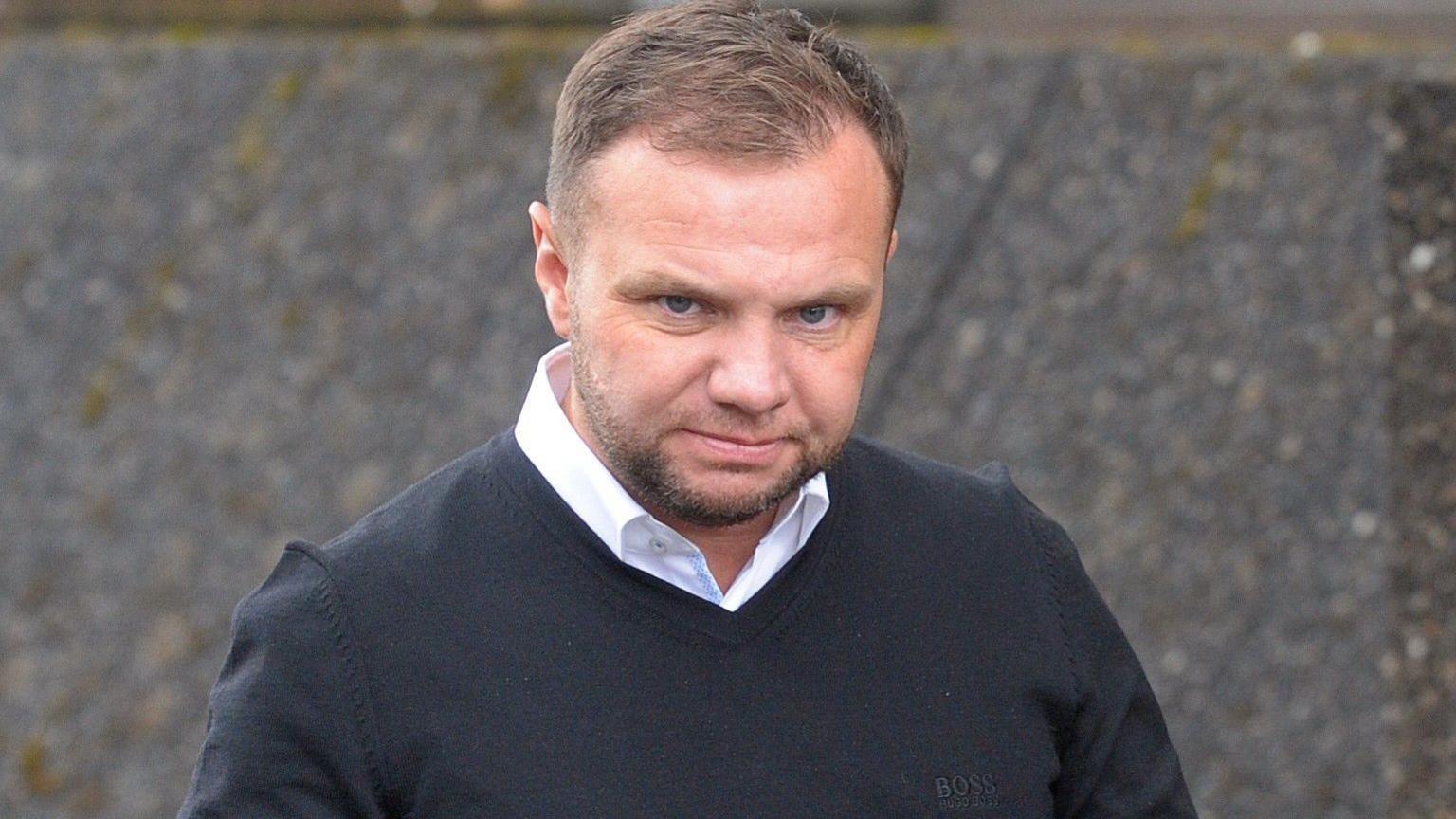 Head and shoulders shot of Eddie Lyons Jnr looking directly at the camera as he walks towards a court building. He has brown hair, combed to the side, and is wearing a black Boss jumper over a white shirt. He has a serious expression and a slightly menacing stare.