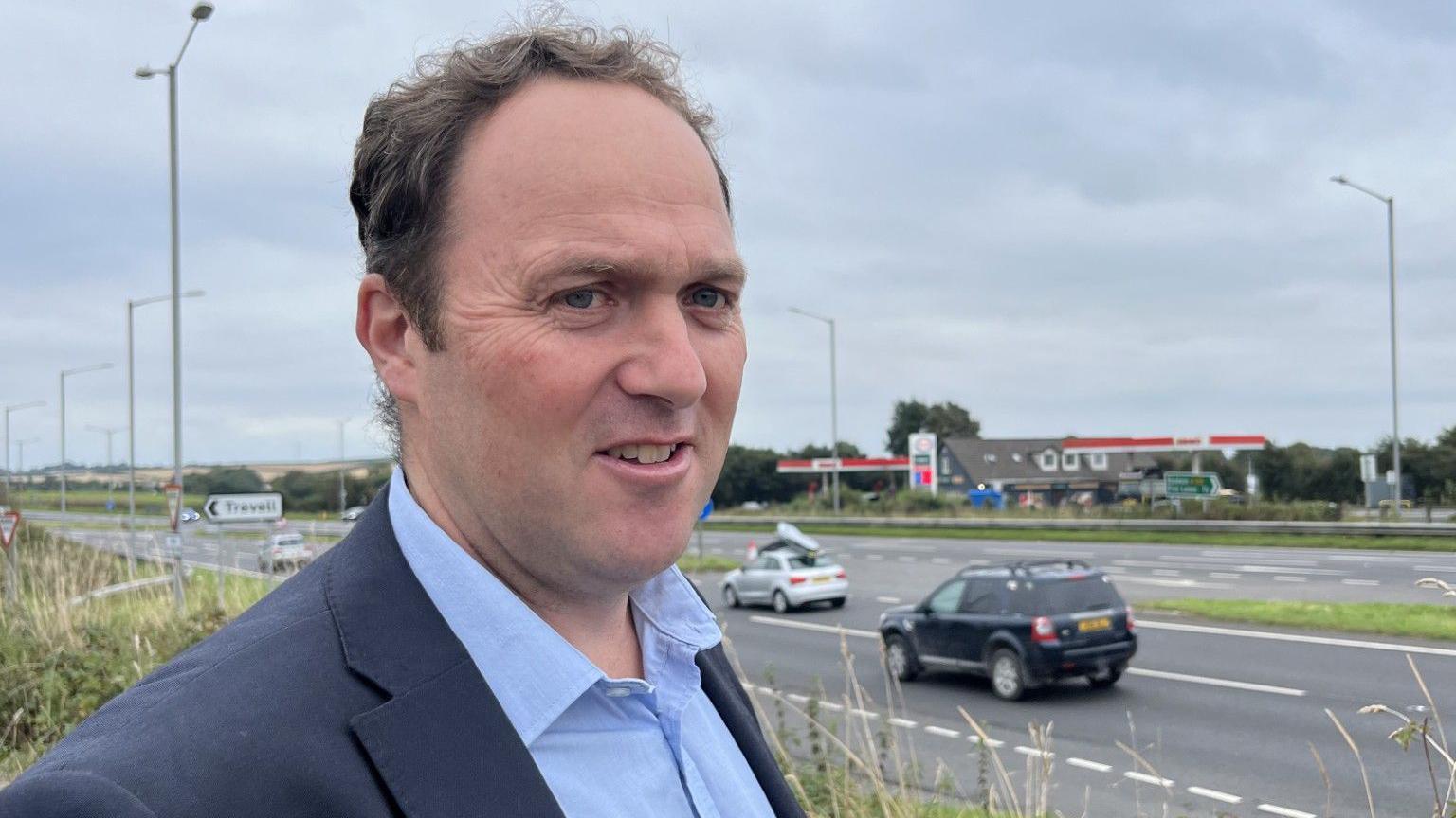 A man in a suit jacket and blue shirt standing beside the A30 at Plusha, with cars and a petrol station in the background