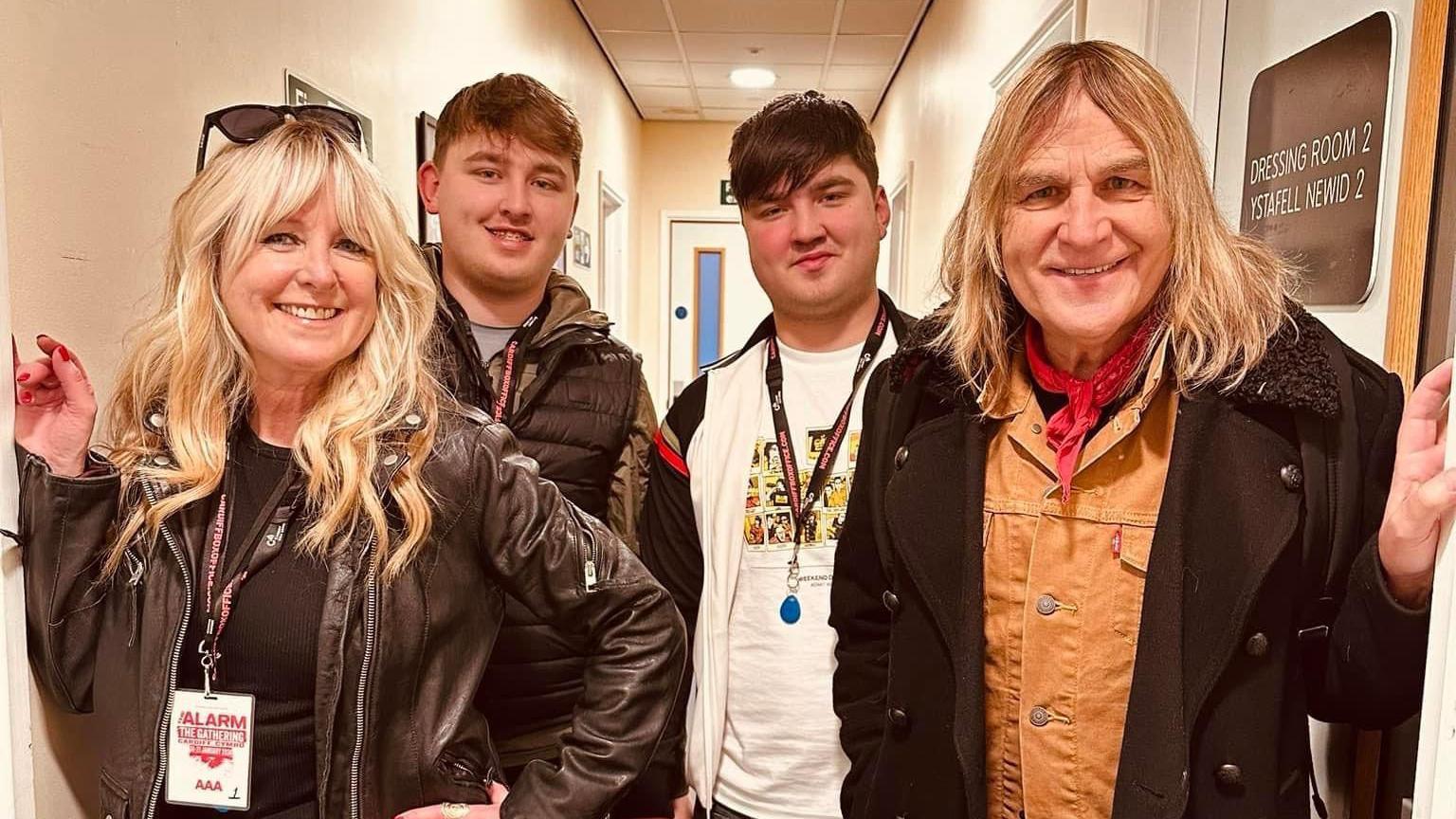 Jules Peters (left) with Mike Peters (right) and their two sons (centre). They all smile towards the camera. They are in a corridor with a dressing room door sign to their right, with Jules and the teenage boys wearing lanyards. Jules is wearing a leather jacket with her long hair in waves, while Mike is wearing an orange denim jacket with a black coat on top, and a red neck tie. 