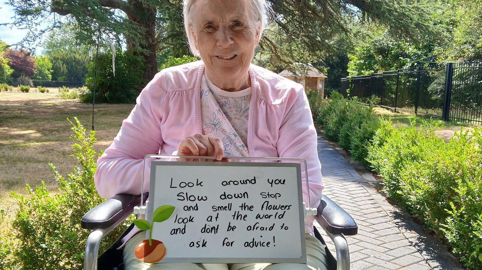 A woman with short grey hair and wearing a pink cardigan, sitting in a wheelchair and holding up a whiteboard saying "look around you, slow down, stop and smell the flowers, look at the world and don't be afraid to ask for advice". It's a sunyn day and there's a garden behind her.