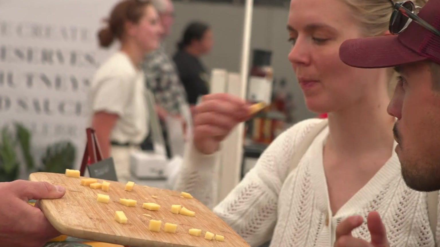 Two people taste small amounts of cheese on a wooden board at a show.