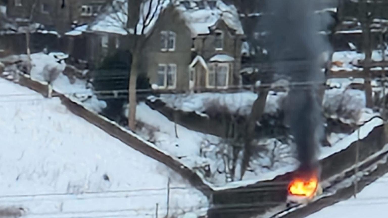 A police van on fire on a snowy road, with the large house in the background.