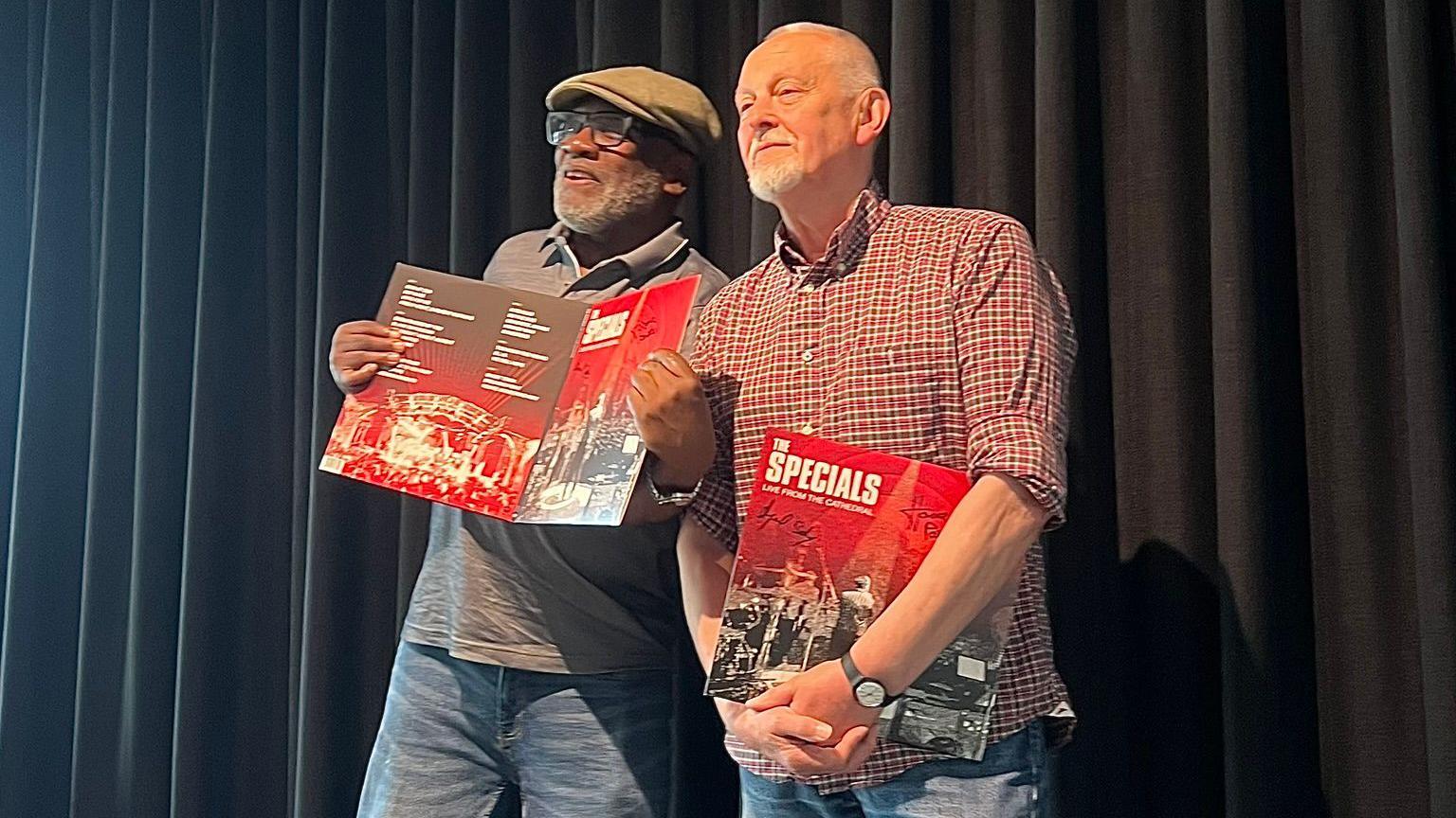 Lynval Golding, wearing a flat cap and glasses, holds up a copy of the new album as he stands in front of a dark curtain. Horace Panter is standing beside him wearing a red checked shirt with a copy under his arm. 
