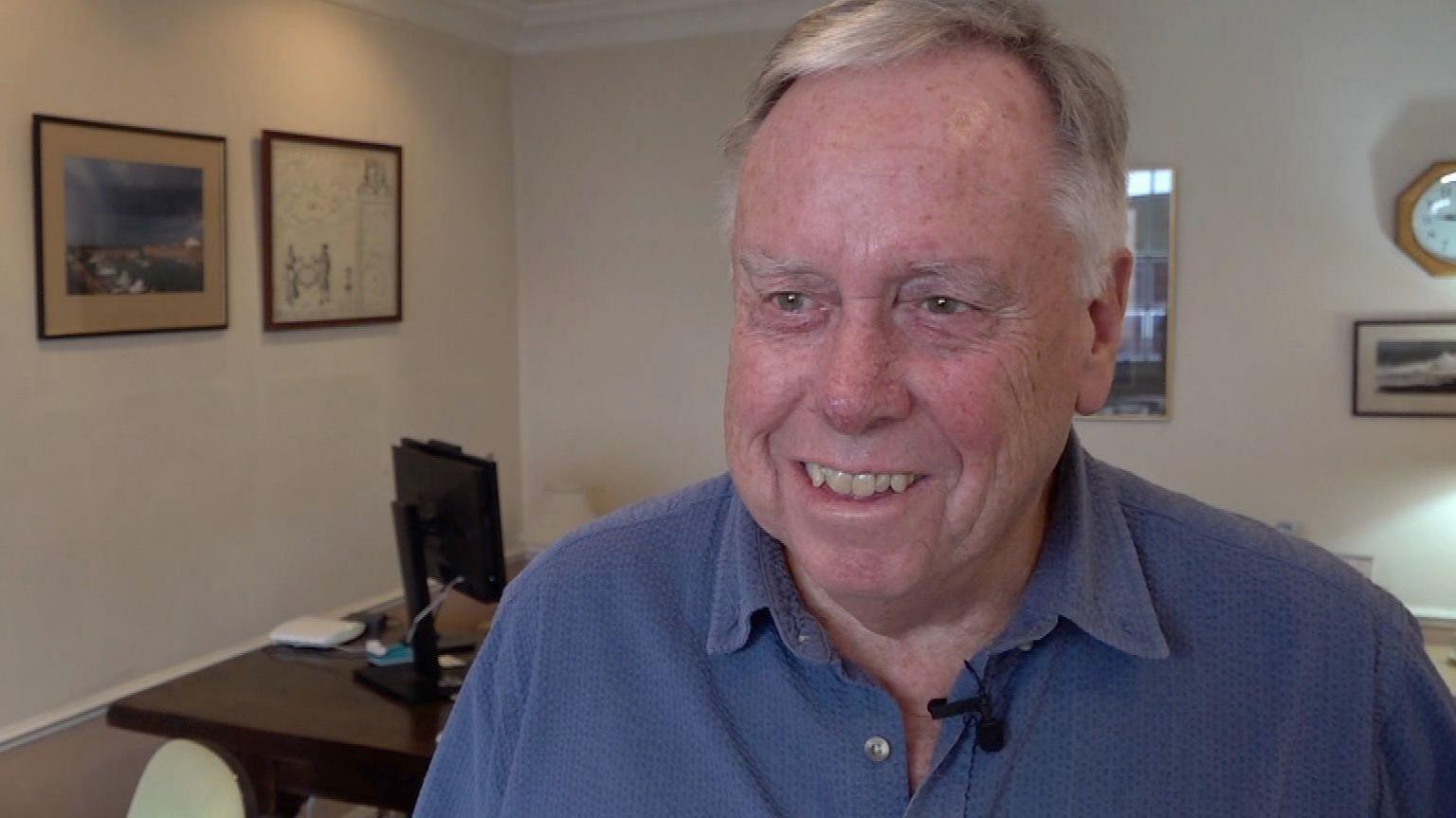 Andrew Eggleston wearing a blue shirt and looking at the camera and smiling. Behind in is a desk with a computer on it. To the left of him is a wall with paintings on. Behind him is a wall with paintings and a clock.  