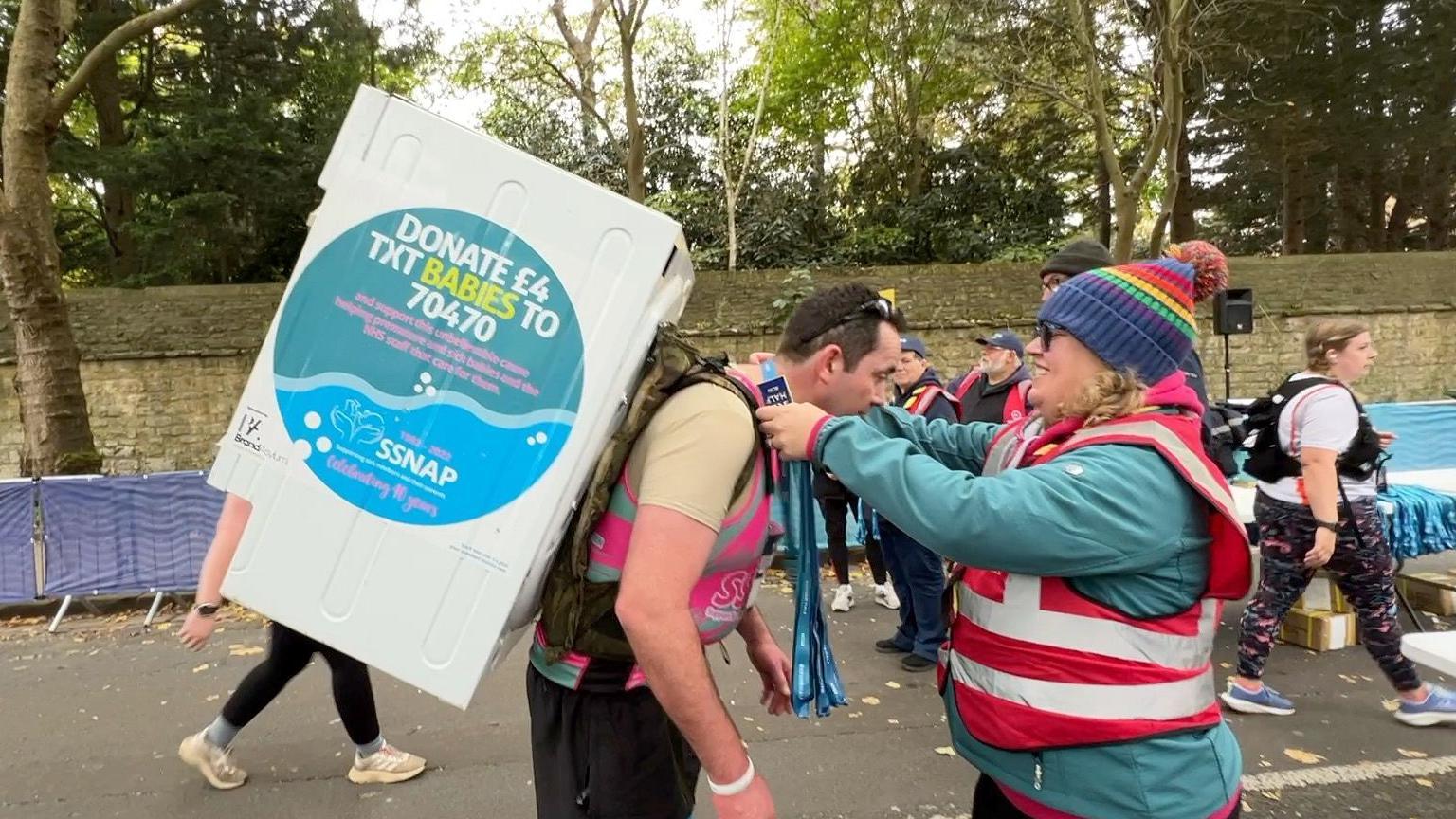 A man wearing a pink vest has a washing machine strapped to his back. A volunteer is placing a medal over his neck.