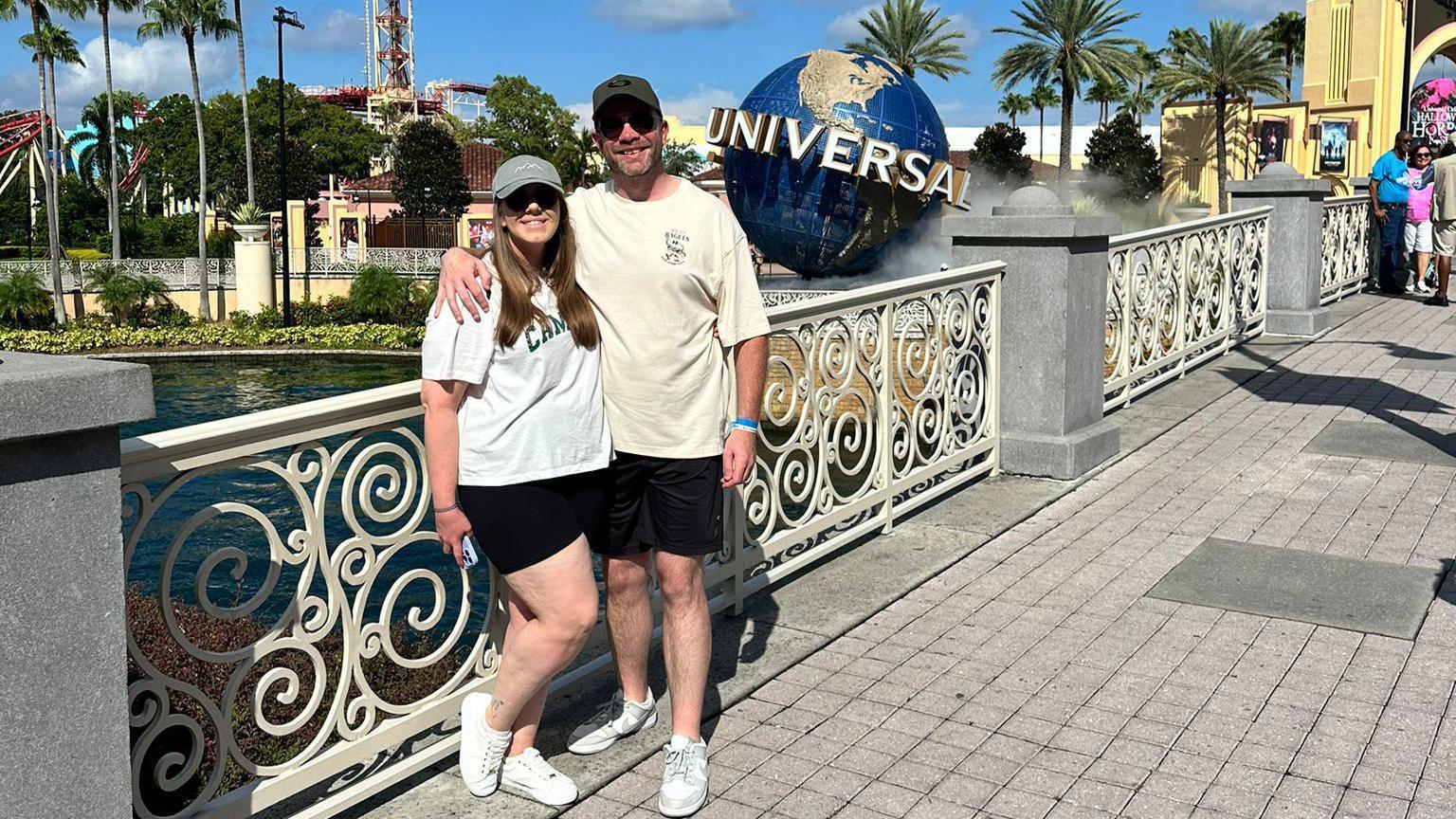 A woman with a white t-shirt, black shorts, white trainers, a grey baseball cap and sunglasses standing next to a man wearing a cream t-shirt, black shorts, white trainers, a black baseball cap and sunglasses against white railings, with the Universal Orlando resort in Florida pictured the background.