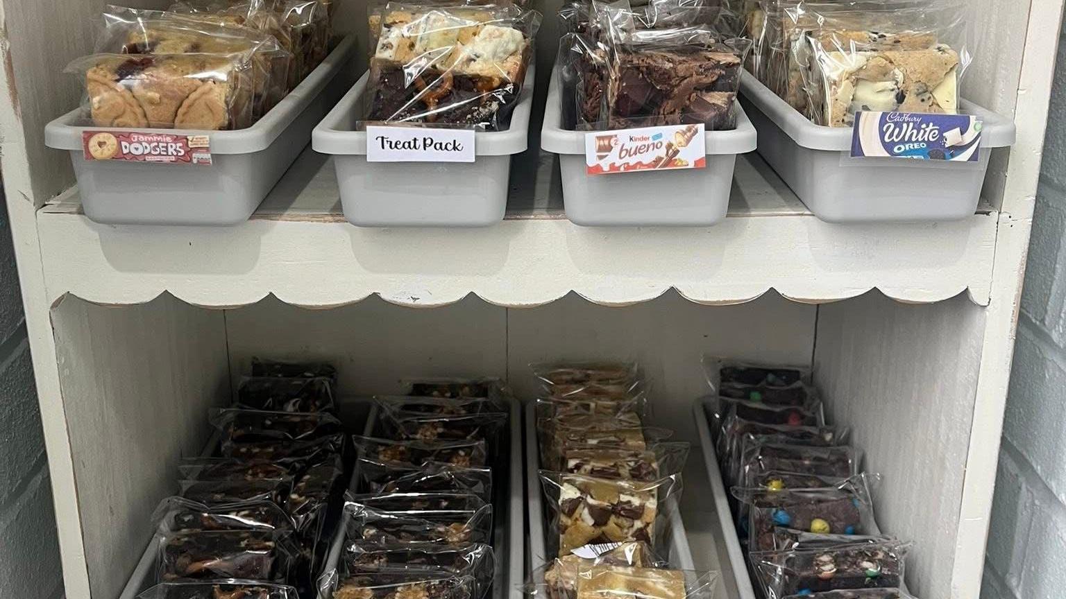 A selection of cakes, in clear plastic packaging, sit in trays across two white shelves. 