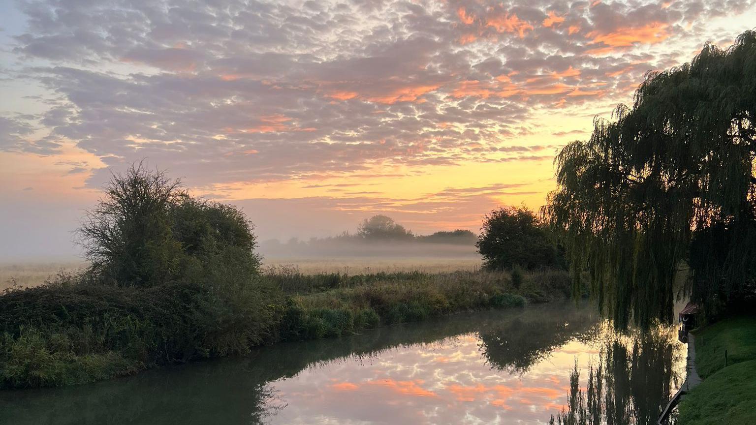 A misty sunrise over a river. There are patchy orange and pink clouds in the sky which are reflected in the water. On either side of the river there are fields and trees.