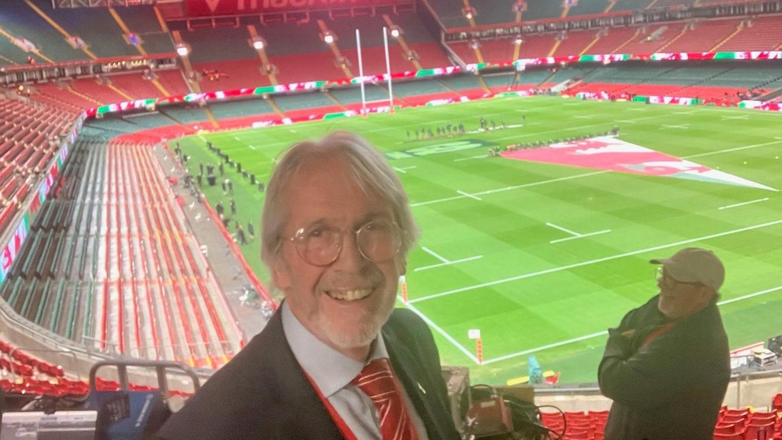 Iestyn Garlick yn gwenu at y camera gyda chae'r stadiwm yn y cefndir a seddi gwag. Mae e'n gwisgo sbectol, tei coch a siwt.