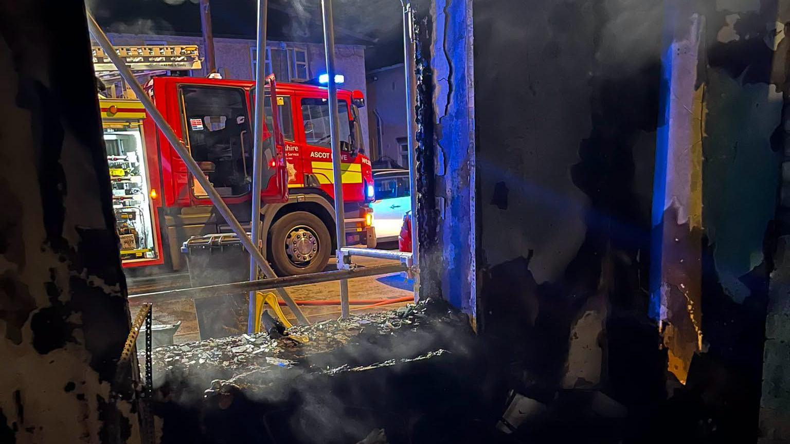 A photograph from the inside of a severely fire damaged house. The window is smashed and outside a fire engine is parked on the road. 