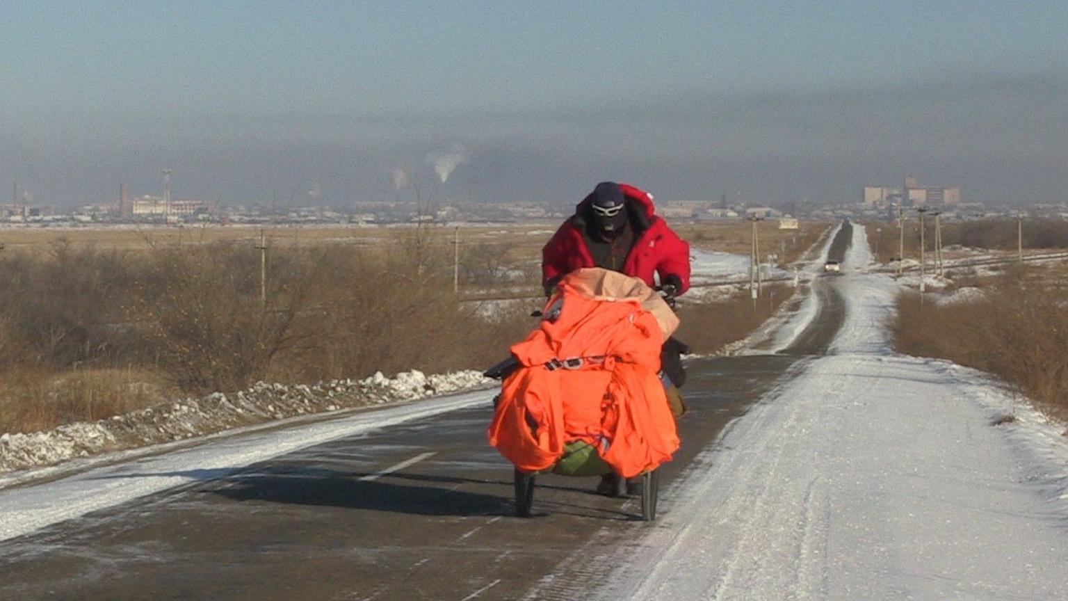 Karl Bushby wearing a balaclava, sunglasses and a red coat pushing a buggy containing his belongings along a snow covered road. 