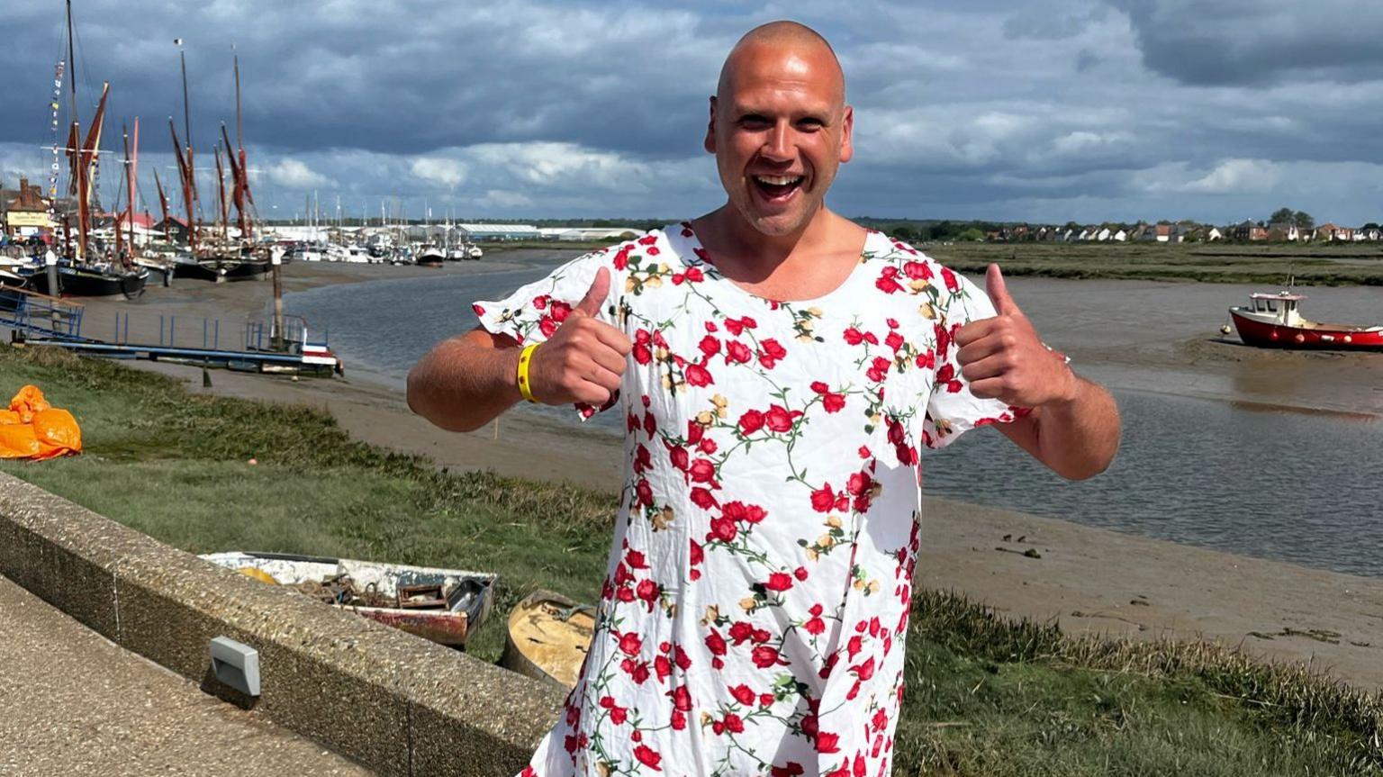A man smiling, dressed in a white dress with red flowers, with his thumbs up