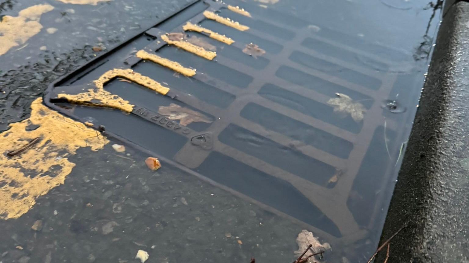 A close up picture of a street grid drain that has water floating on top of it, it is clearly overwhelmed. There is a kerb next to it and yellow lines painted over part of the drain.
