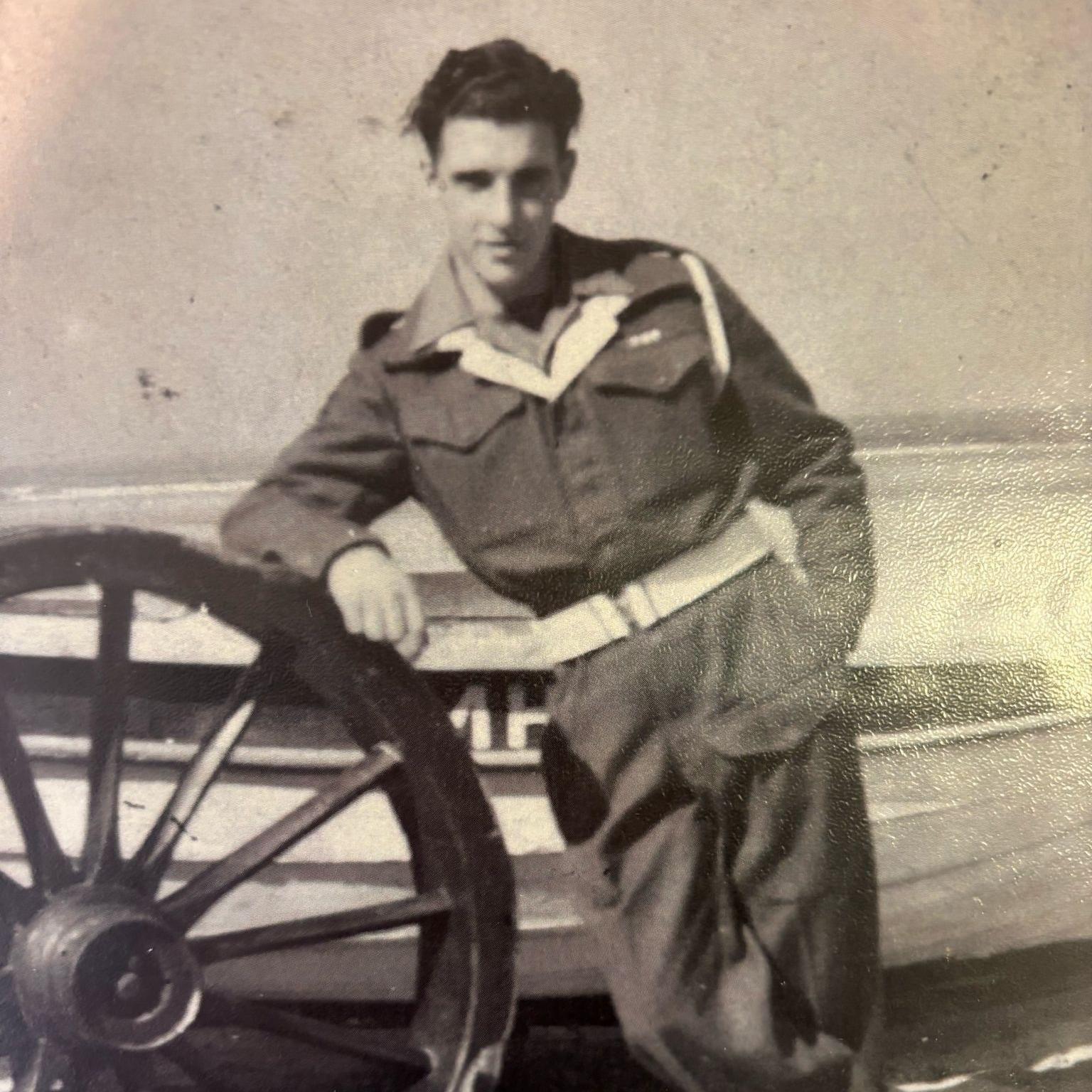 A black and white photo of Dennis Smith leaning on a large wheel in his military uniform. He has short brown hair. Behind him is a small white boat.