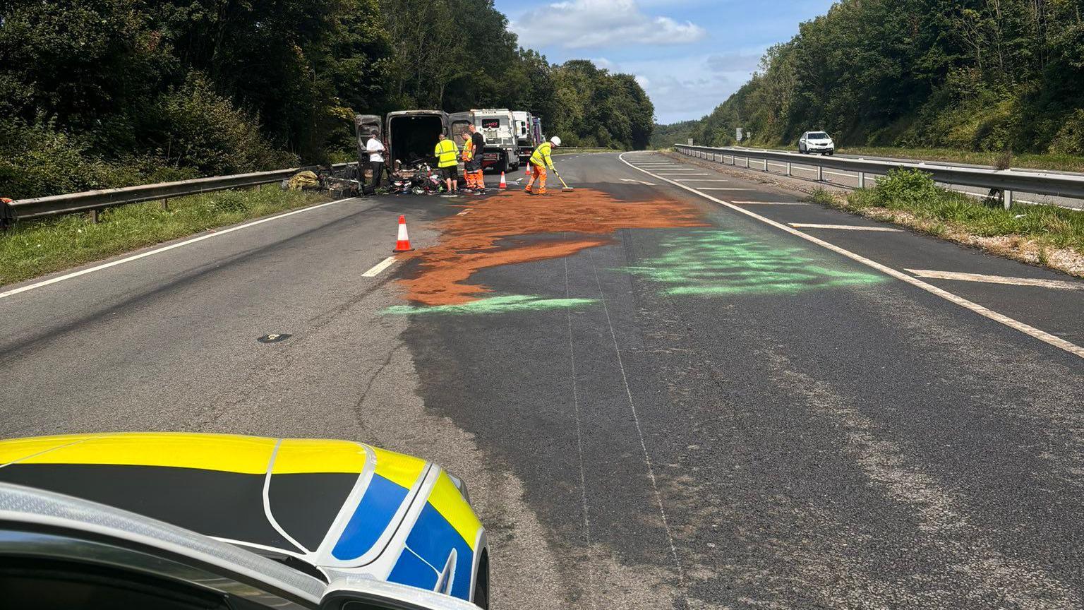 Van fire on A380, with brown and green colours on carriageway and workers in the background near burnt-out vehicle.