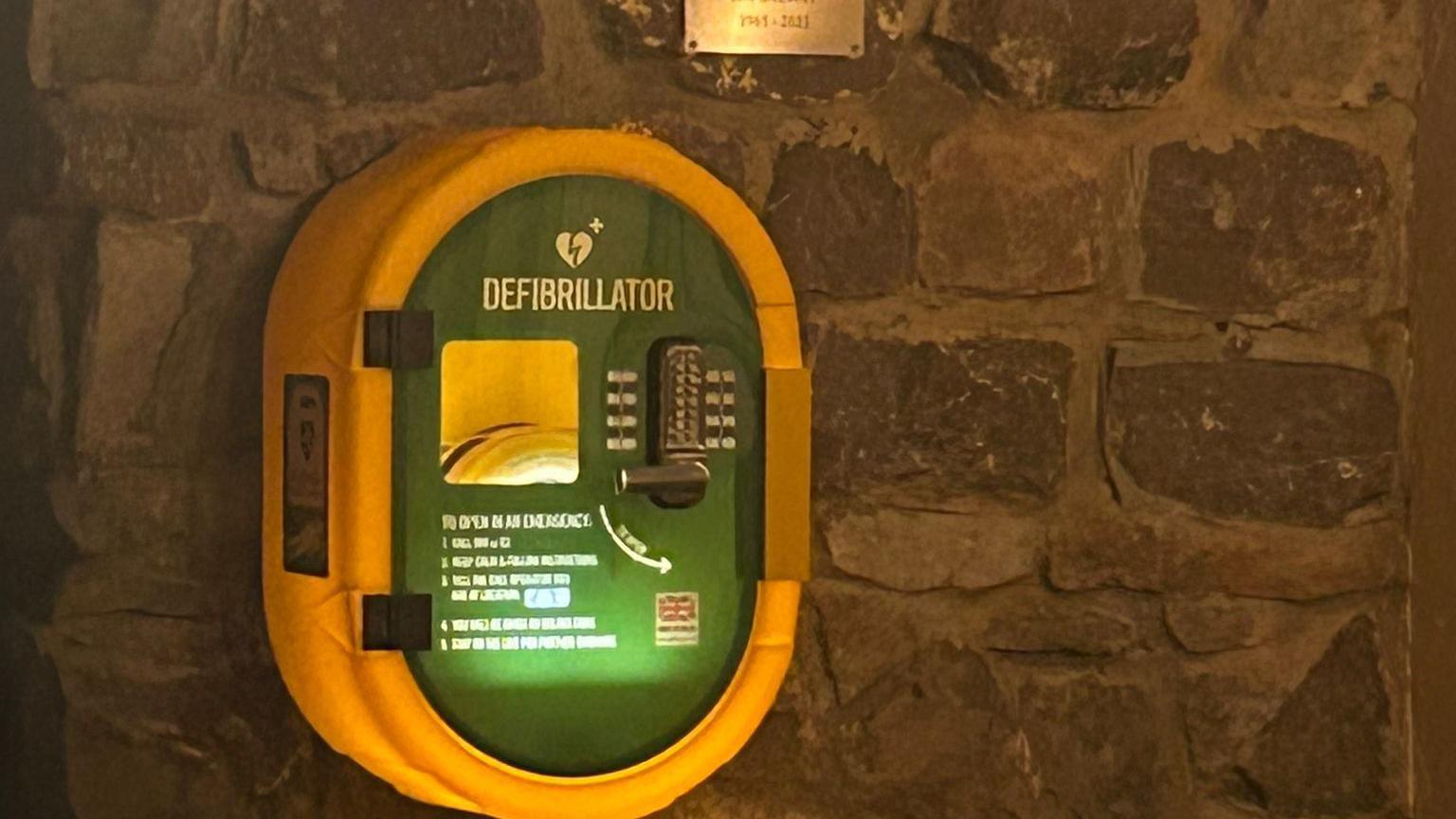 A green and yellow defibrillator installed on a stone wall. It has a keypad lock and the instructions are lit up. 