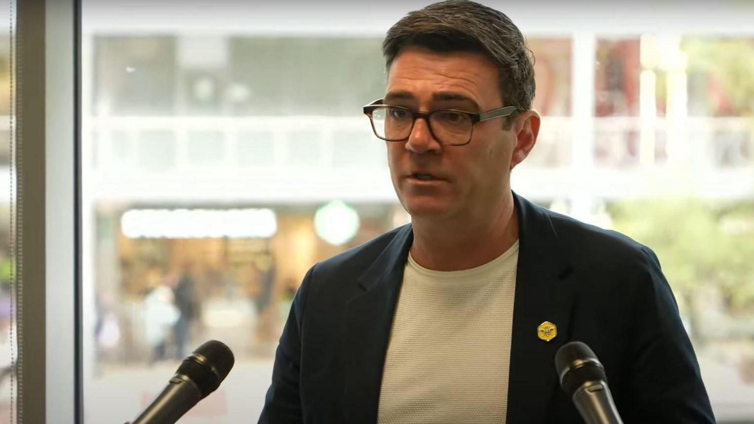 Andy Burnham wearing a white t-shirt and blazer with a Bee Network yellow badge addresses a press conference from a podium.
