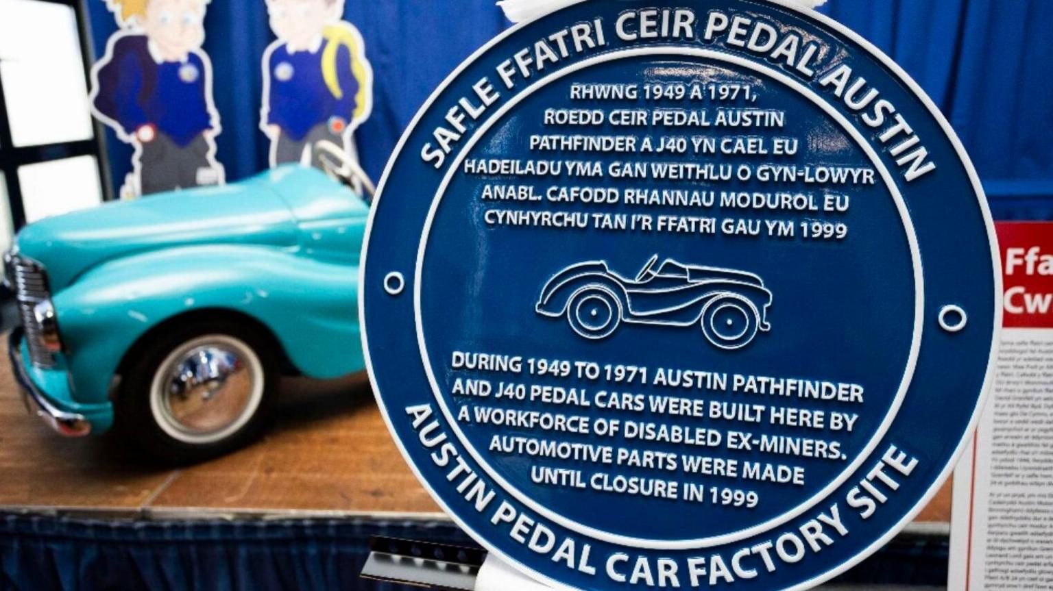 A blue plaque with white writing. It shows a line drawing of a pedal car, and it bears the inscription, in Welsh and English: Austin Pedal Car Factory site.
During 1949 to 1971 Austin Pathfinder and J40 pedal cars were built here by a workforce of disabled ex-miners. Automotive parts were made until closure in 1999.