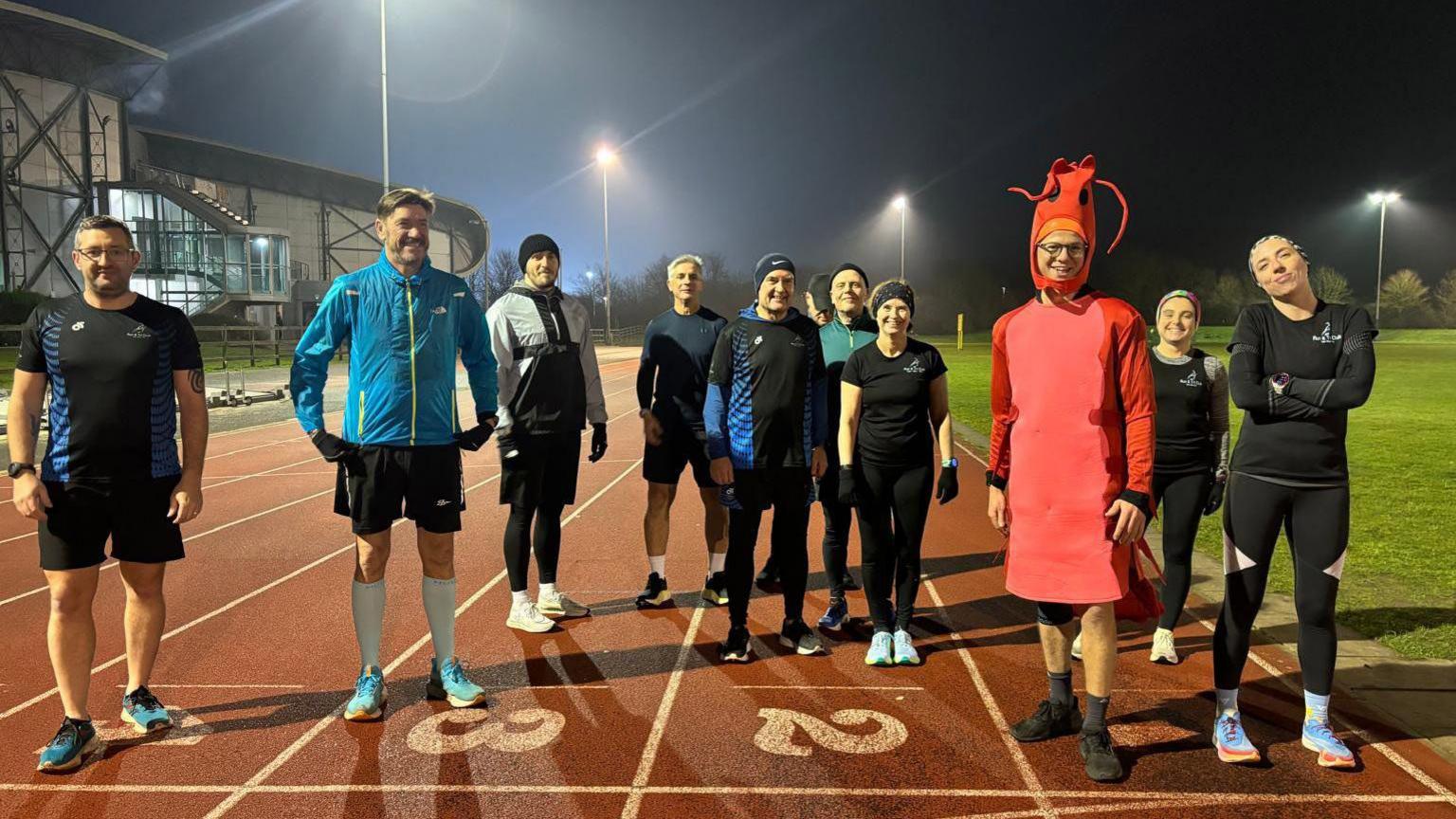 Jamie is standing on the right of the picture wearing the shrimp costume. There are nine people crowded around him, standing on a running track. They are all smiling towards the camera. 