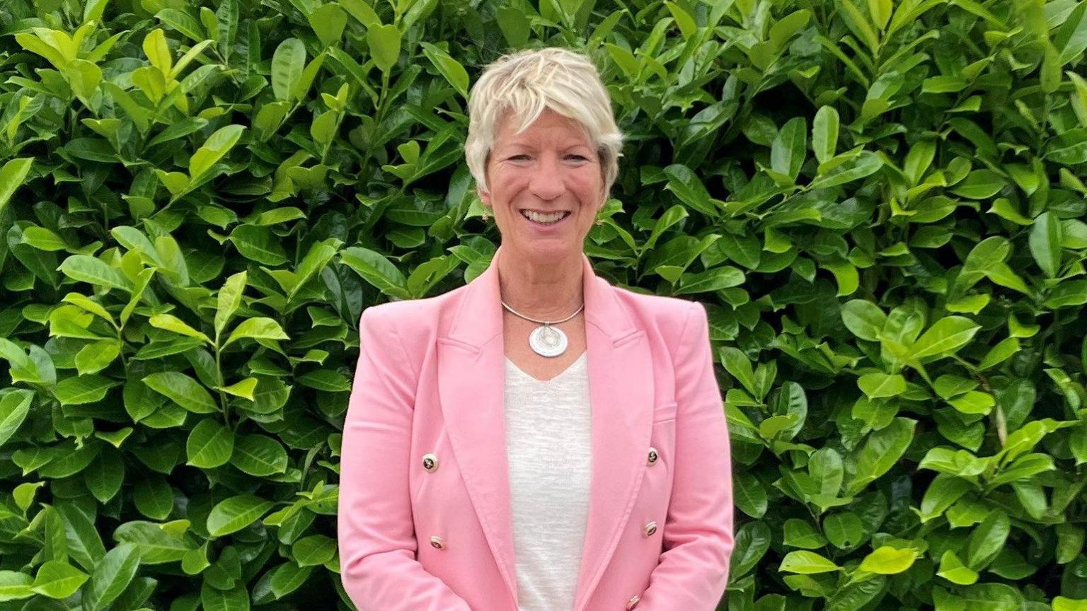 Pippa Heylings wearing a pink suit and grey top, standing in front of a green hedge