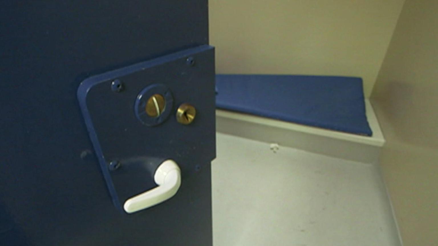 Close up shot of a blue custody suite door which is slightly open, showing the inside of the cell. A blue mat is on a raised section of the cell. The door handle is white and a set of locks are on the door.