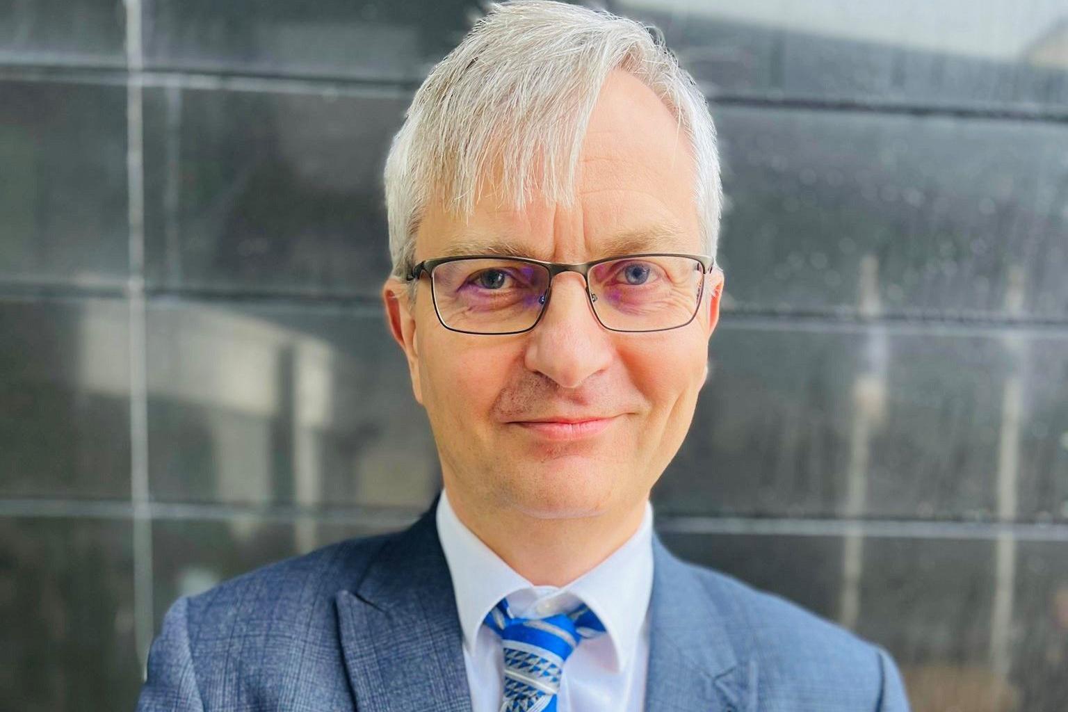 Man with grey hair and glasses, wearing a white shirt, blue striped tie and light blue checked suit jacket, smiling and looking at camera.