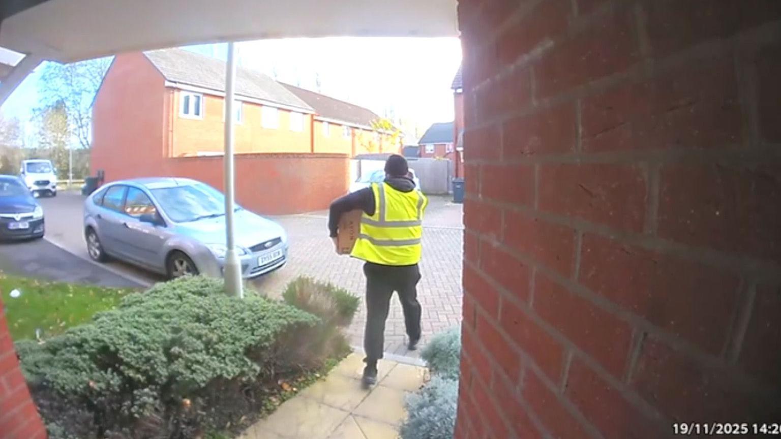 A picture taken from a doorbell camera showing a man wearing a high-viz jacket and a black hat walking away from a house with a parcel under his arm.