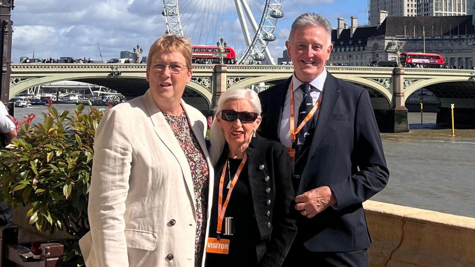 Three people are pictured next to the River Thames in London with the London Eye in the background - a woman with short blonde hair and glasses is wearing a beige blazer and patterned dress.  She is next to a woman with white hair, sunglasses and wearing black.  A man is also picture wearing a suit and smiling. Behind them is a bridge and the river Thames and also the London Eye.