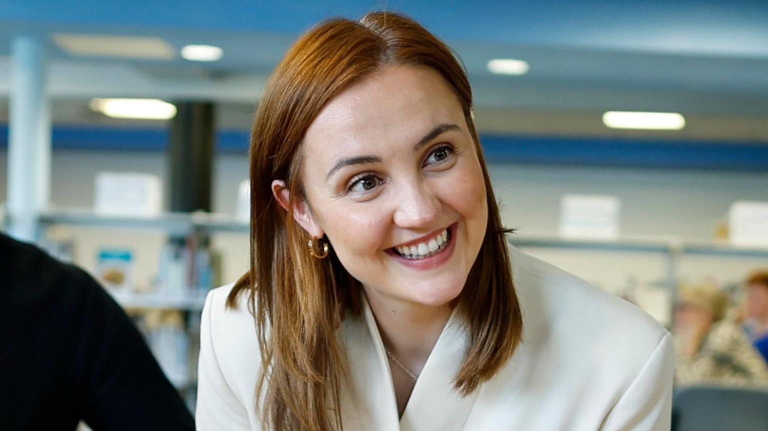 Head and shoulders shot of a smiling Mairi McAllan in a classroom setting. She is wearing a white jacked and has long brown hair.