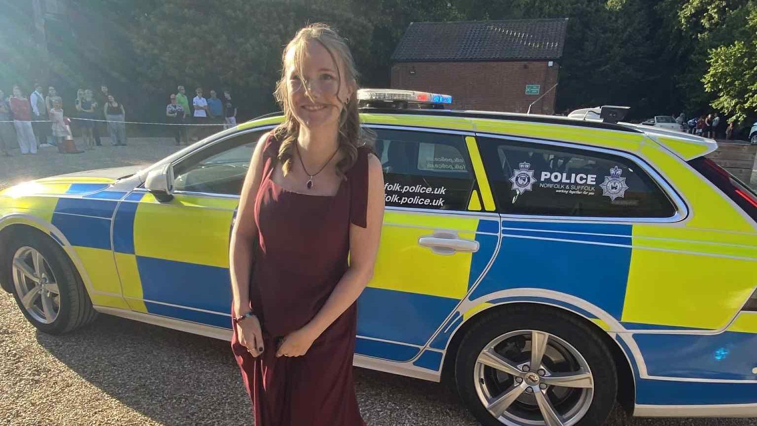 Evie stands in front of a Suffolk Police car at her prom. She has brown hair that is half tied back and half down and slightly curled. 