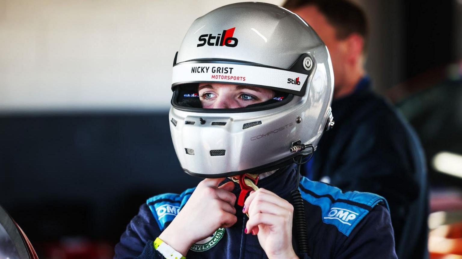 Daniella is wearing a silver racing helmet and a dark blue racing suit. She is fixing the strap on helmet and looking away from the camera.