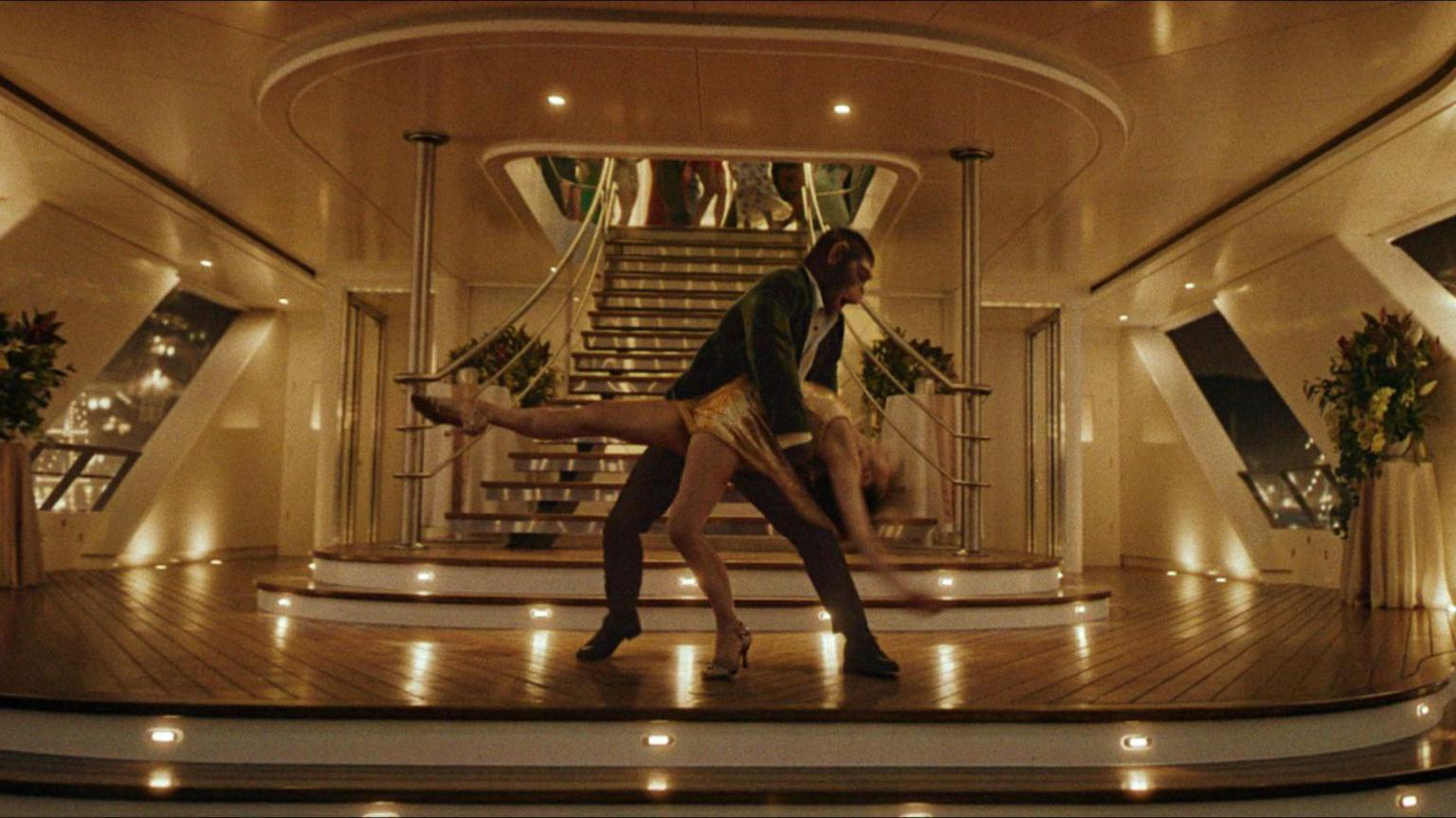 A human-sized chimpanzee wearing a suit is ballroom dancing with a woman on a lower level of yacht with a party upstairs at night time.