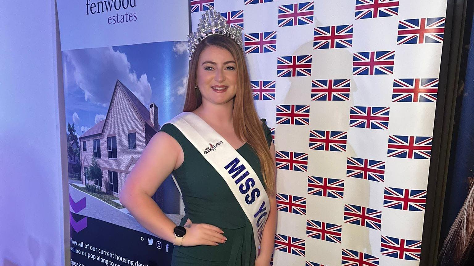 A woman with long brown hair, wearing a green dress, crown and a sash which says 'Miss York 2025', is posing for a photo. She has one hand on her hip and is smiling at the camera.