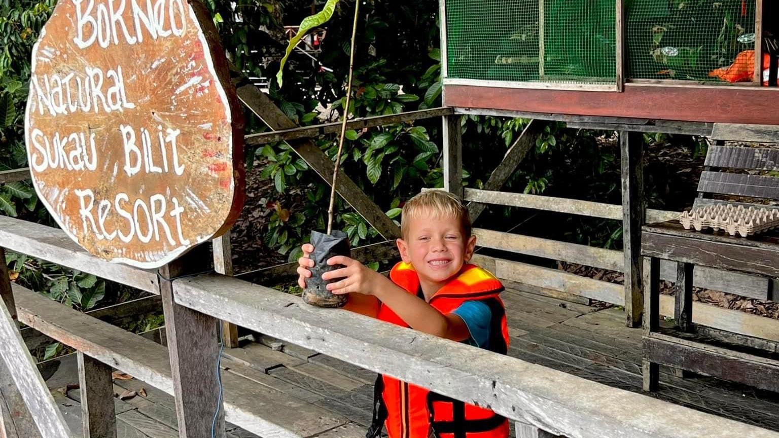 Jamie with blond hair wearing am orange gillet and blue t-shirt smiling as he plants a tree in Borneo standing on a wooden structure with shrubs in the background