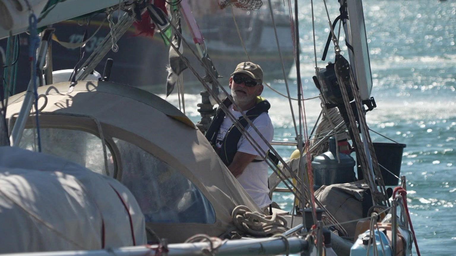 The picture shows a person standing on a sailing boat, wearing a cap and a life jacket. They're positioned near the cockpit area, surrounded by sailing equipment including ropes and sails. In the background, you can see water and a few other boats in the distance.
