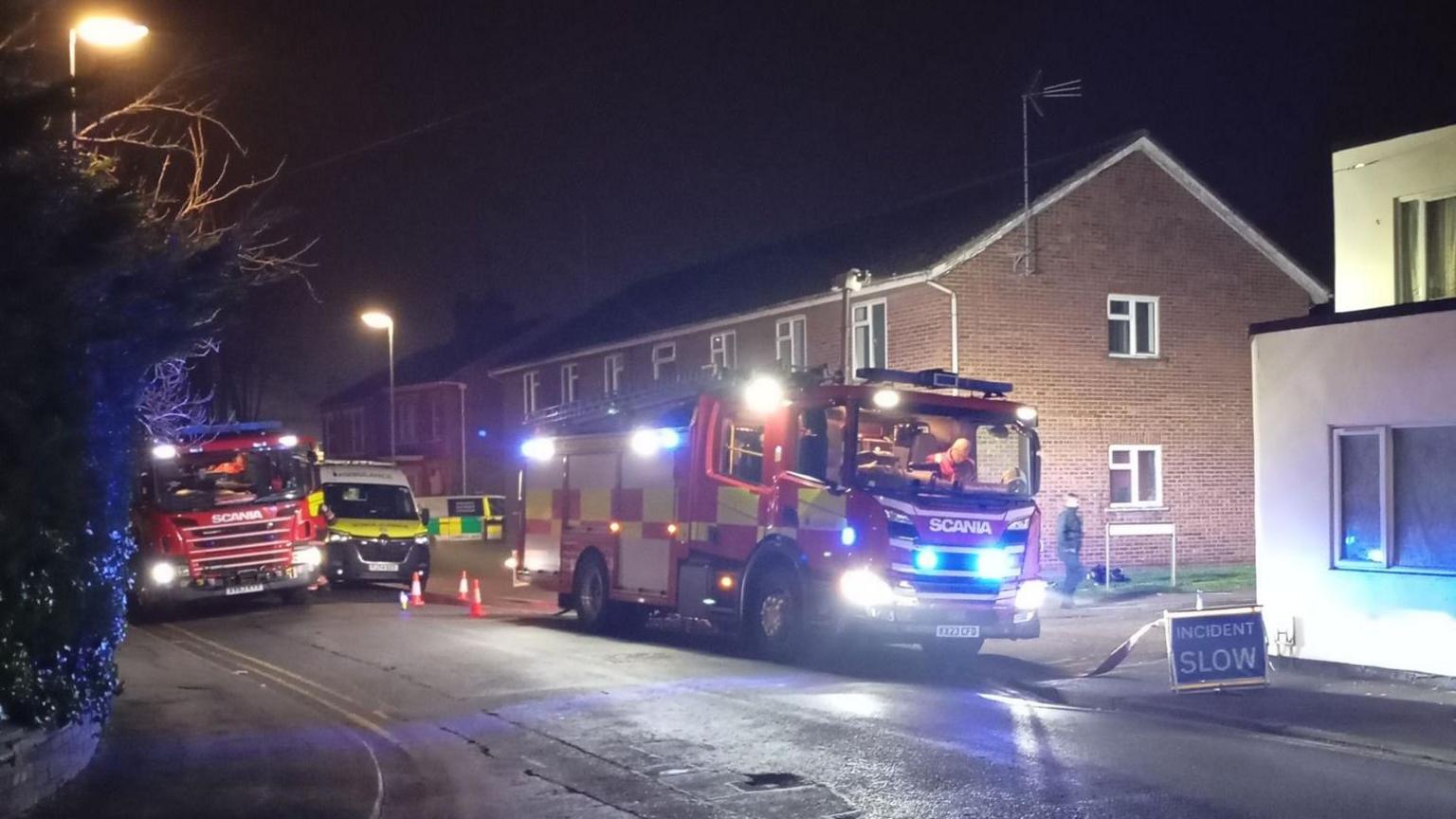 Two fire engines parked on a residential road. Behind the one on the right can be seen an ambulance and behind that another emergency vehicle. It is night and the street lights are on. There is a sign in front of the right engine saying incident slow in white letters on a blue.