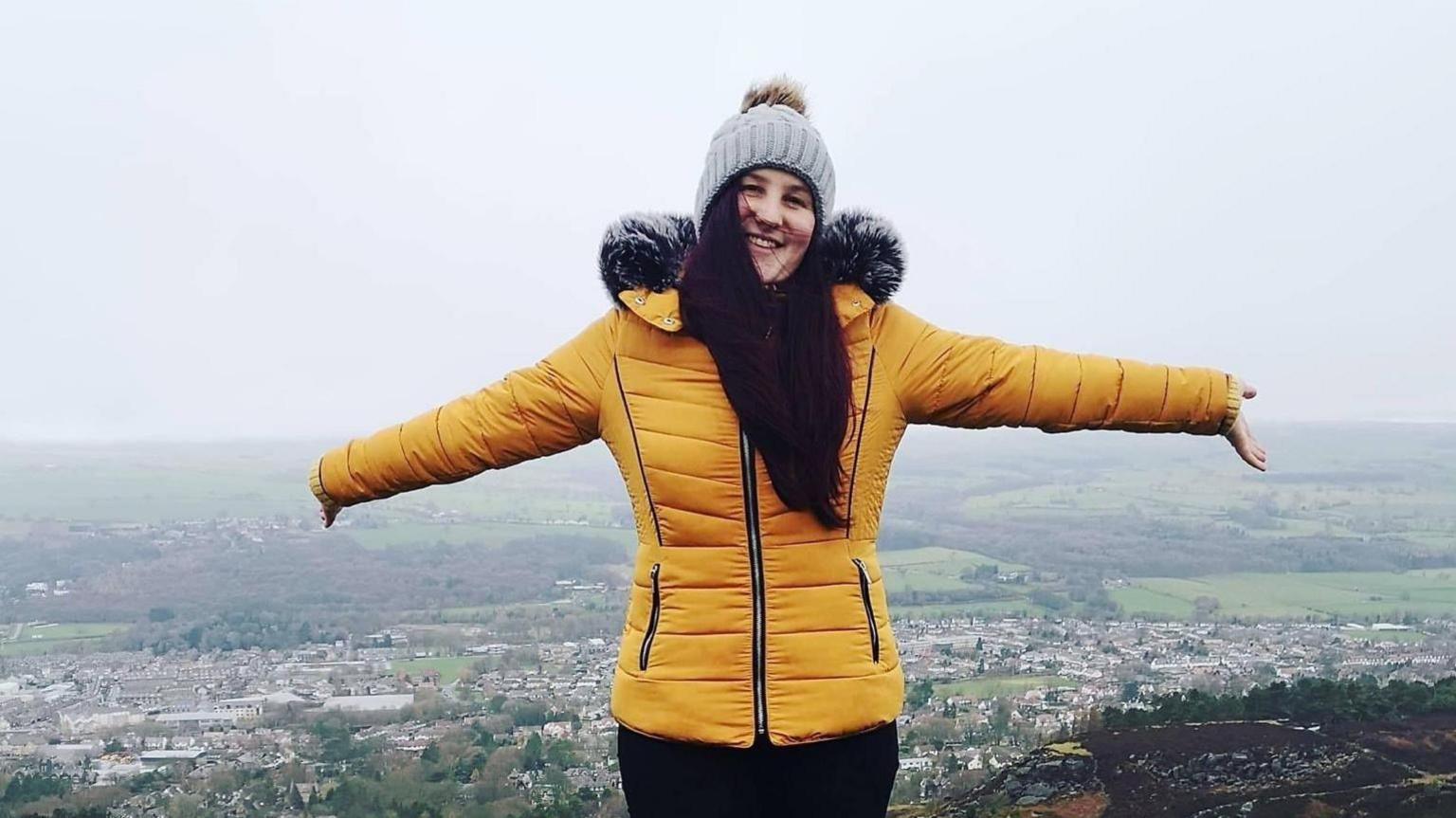 A young woman in her late twenties, standing atop a hill with her arms outstretched either side of her. She is wearing a yellow coat and grey bobble hat.