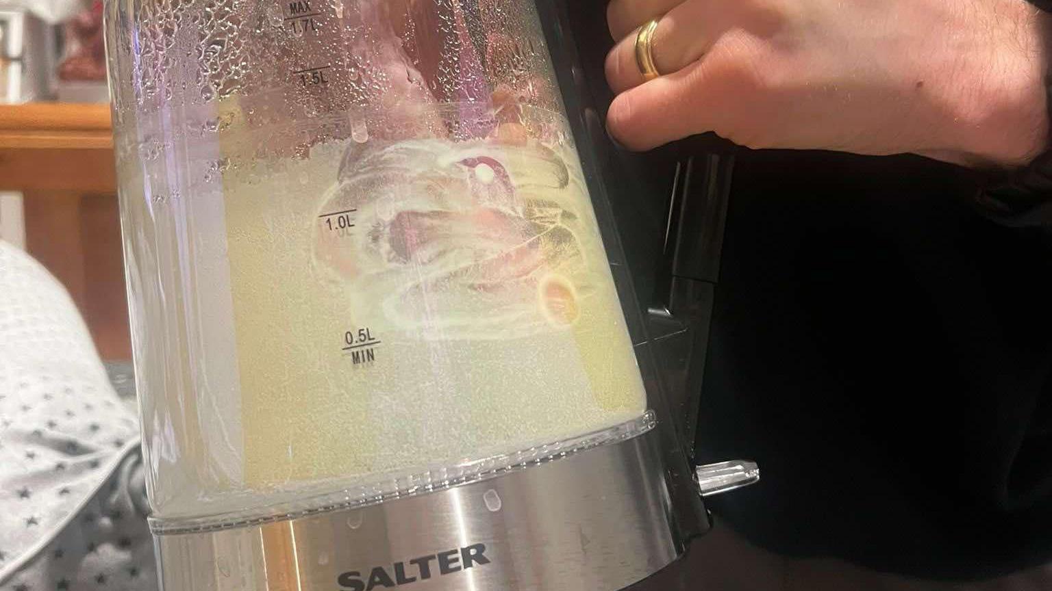 A see through plastic kettle is held up to show how the inside is caked with limescale. The person holding it has their hand inside and is smearing the thick deposit of limescale with their finger.