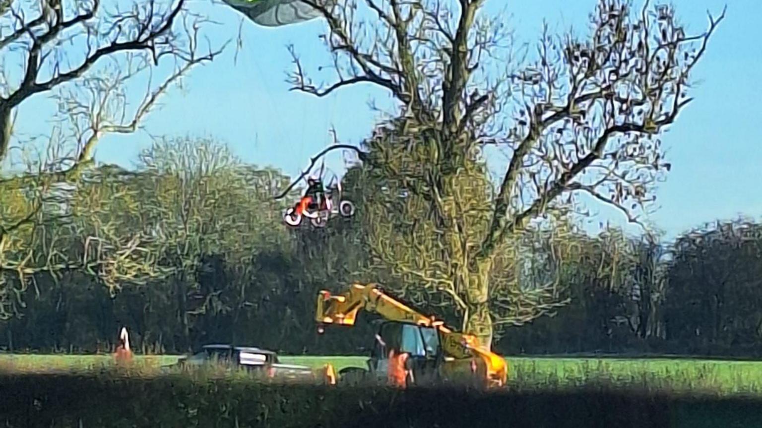 A glider stuck in a tree, with a yellow crane below. There is a man in the glider.