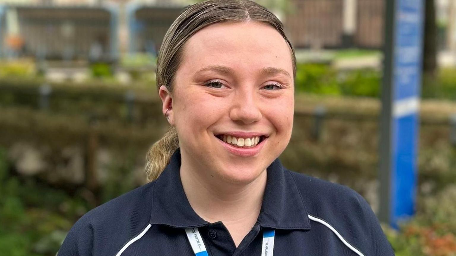 Sian Leader wearing a dark blue medical uniform. Her mid-brown hair is pulled back from her forehead and tied into a pony tail. She is smiling at the camera. 