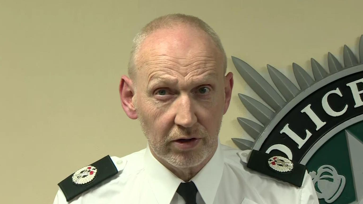 A man with short grey hair and a short beard is talking to someone off camera. He is wearing a white shirt, black tie and has badges on his shoulder. In the background you can see the corner of the PSNI badge on a pale wall. 