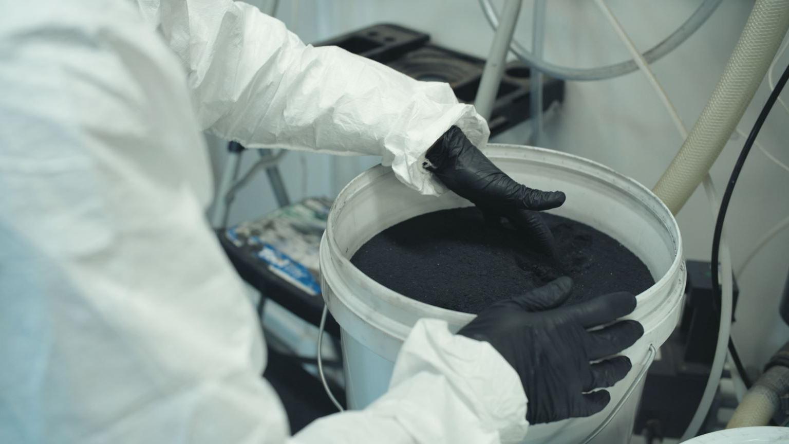 A white bucket containing a black substance with a person in white overalls and black gloves holding the rim
