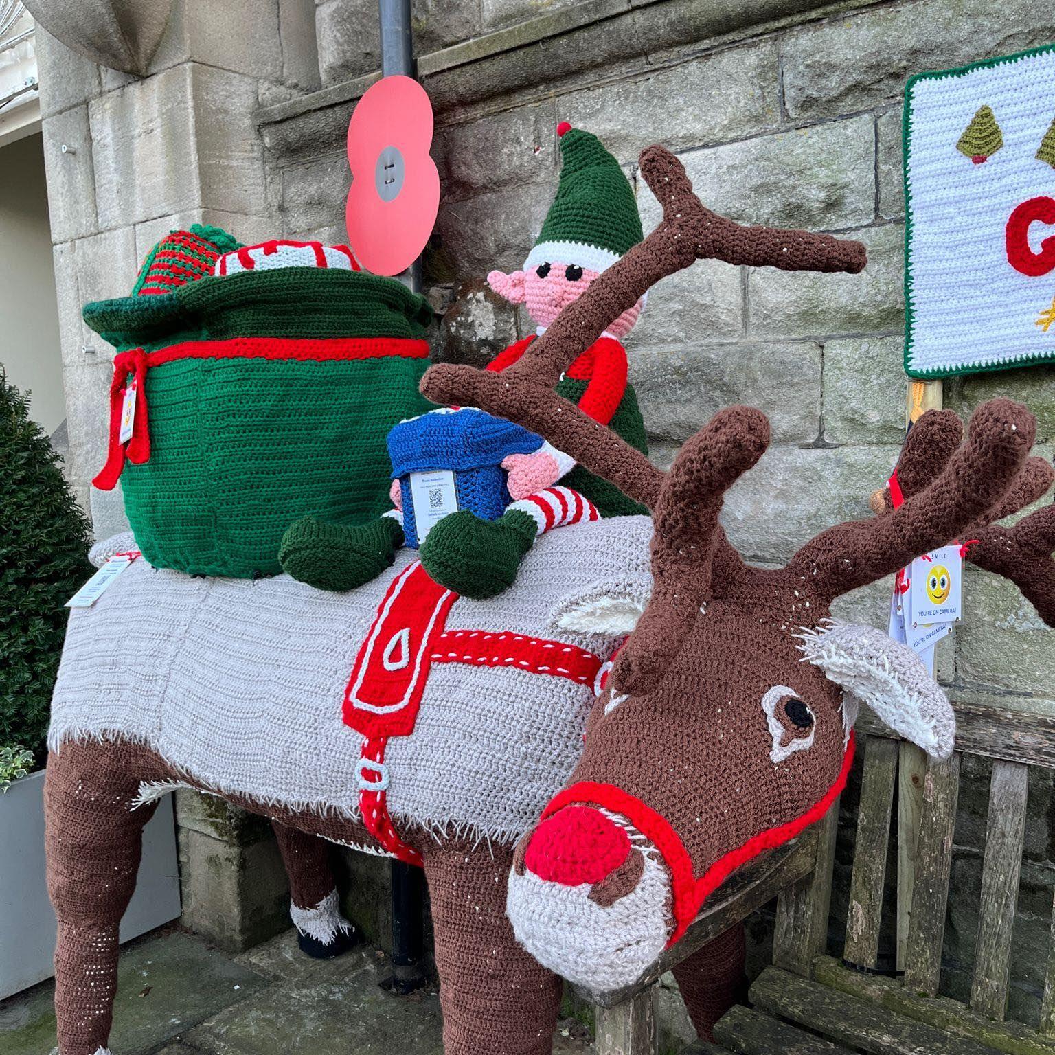 A knitted reindeer faces the camera, with a knitted elf and sack of toys on its back