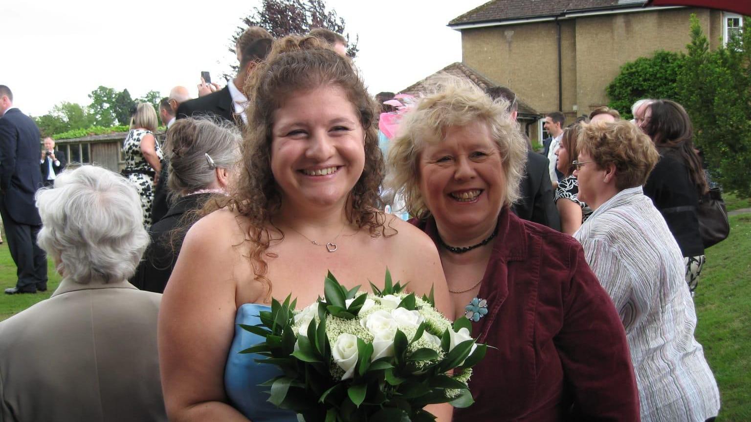 Liz is on the left holding a bouquet and wearing a strapless blue dress.  She has brown, curly hair in a half up-do. Her mother has blonde hair and wears a maroon jacket. 