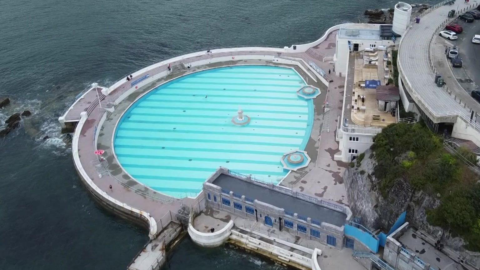 Drone shot of Tinside Lido in Plymouth. The base of the pool is light and dark blue stripes. An area of water called Plymouth Sound is to the left of the lido. There is a road above the lido with some cars parked on it