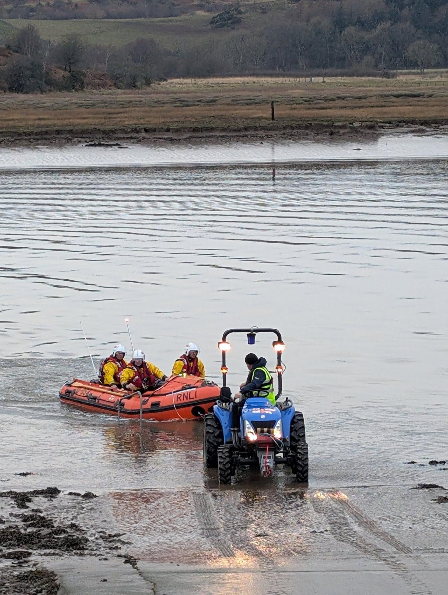 A lifeboat with three crew on board is towed to shore by a small blue tractor coming out of shallow waters on the Solway coast