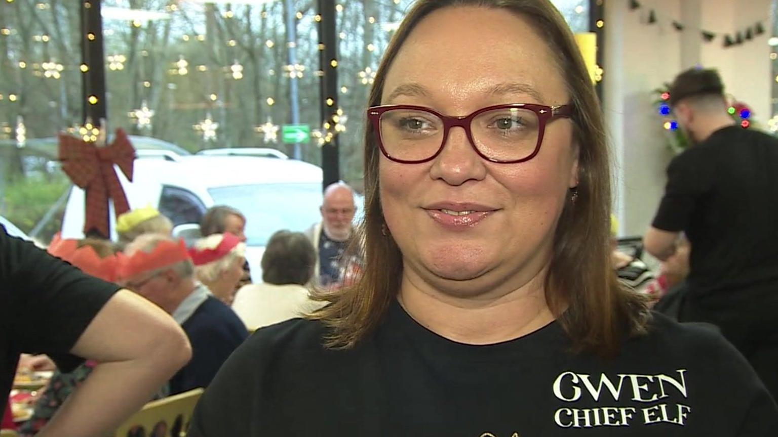 Gwen Geay is smiling at the camera and has brown hair in a bob. She is wearing a black T-shirt that says "Gwen Chief Elf". She has red rimmed glasses on.