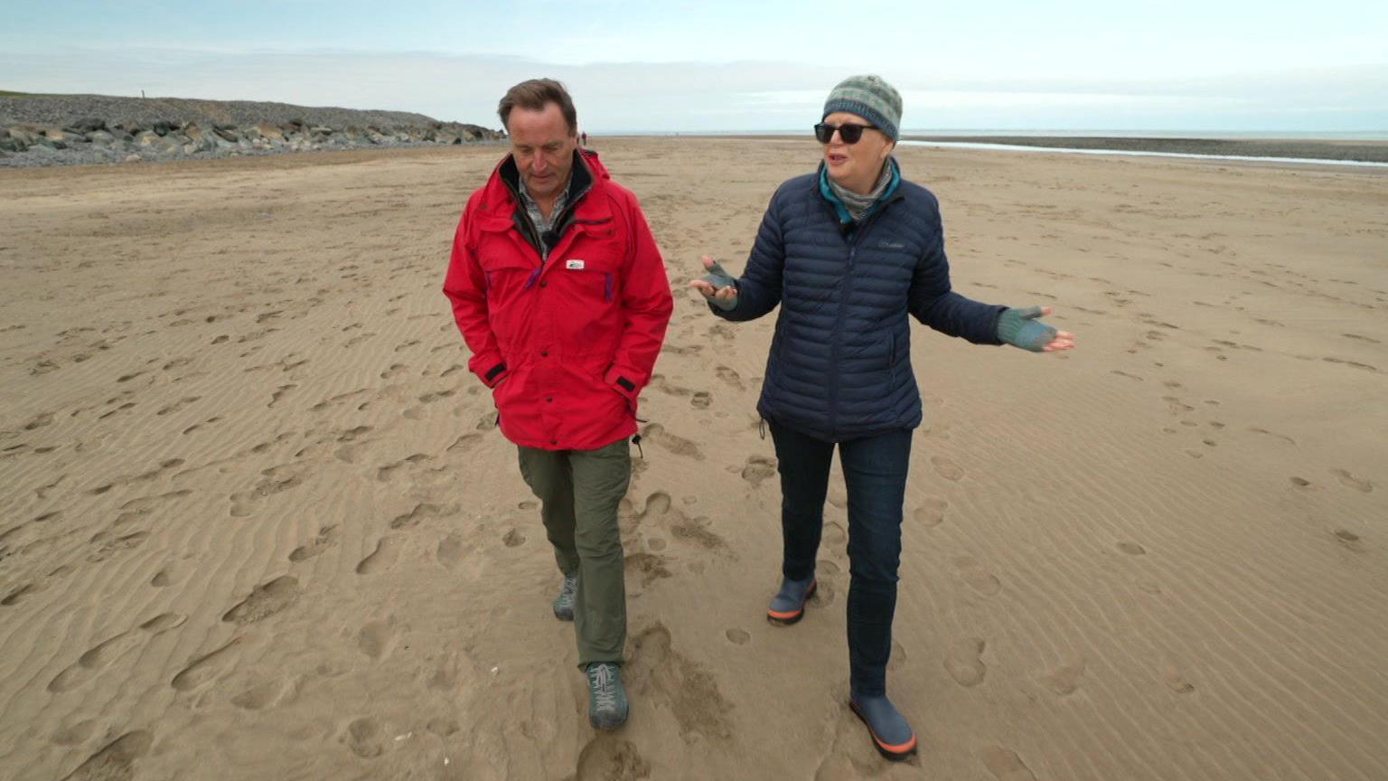 Wrapped up in wintry gloves David Braine is on the left of the shot as Liz talks to him about her love of photography.  They are walking on a sandy beach and footprints can be seen near them.